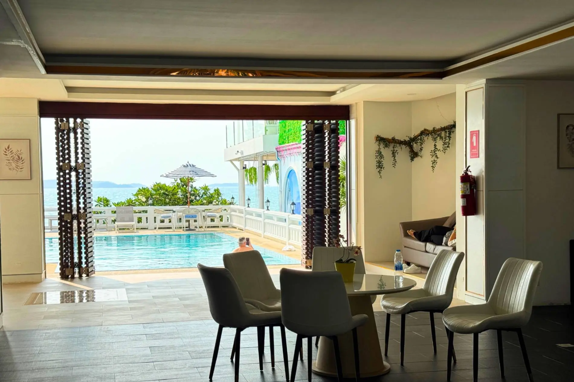 Indoor seating area with white chairs and a table overlooking an outdoor swimming pool and ocean view through open doors.