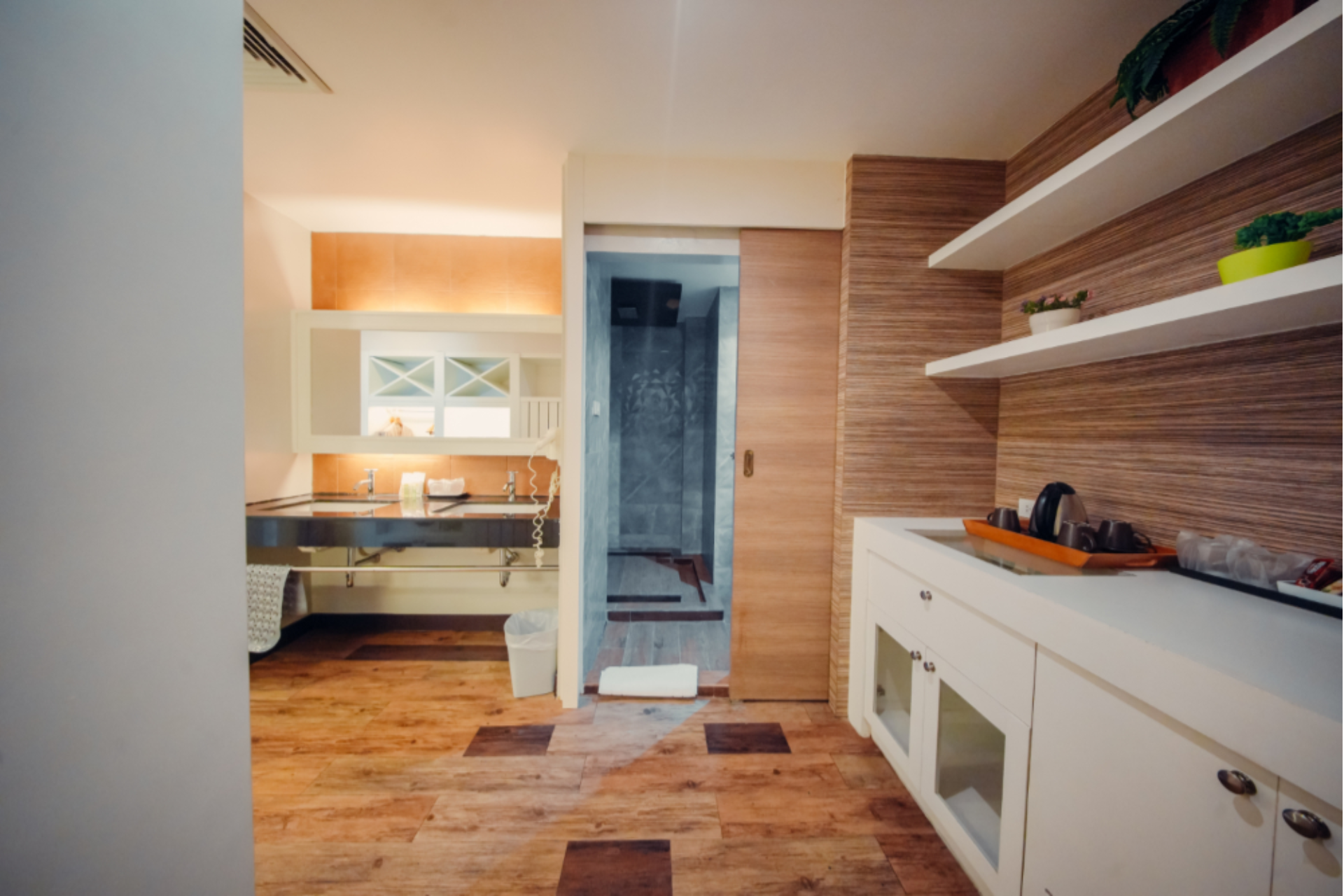 Hotel bathroom with wooden flooring, double sink vanity, open shelves with plants, countertop with kettle and cups, and a sliding door leading to a shower area.