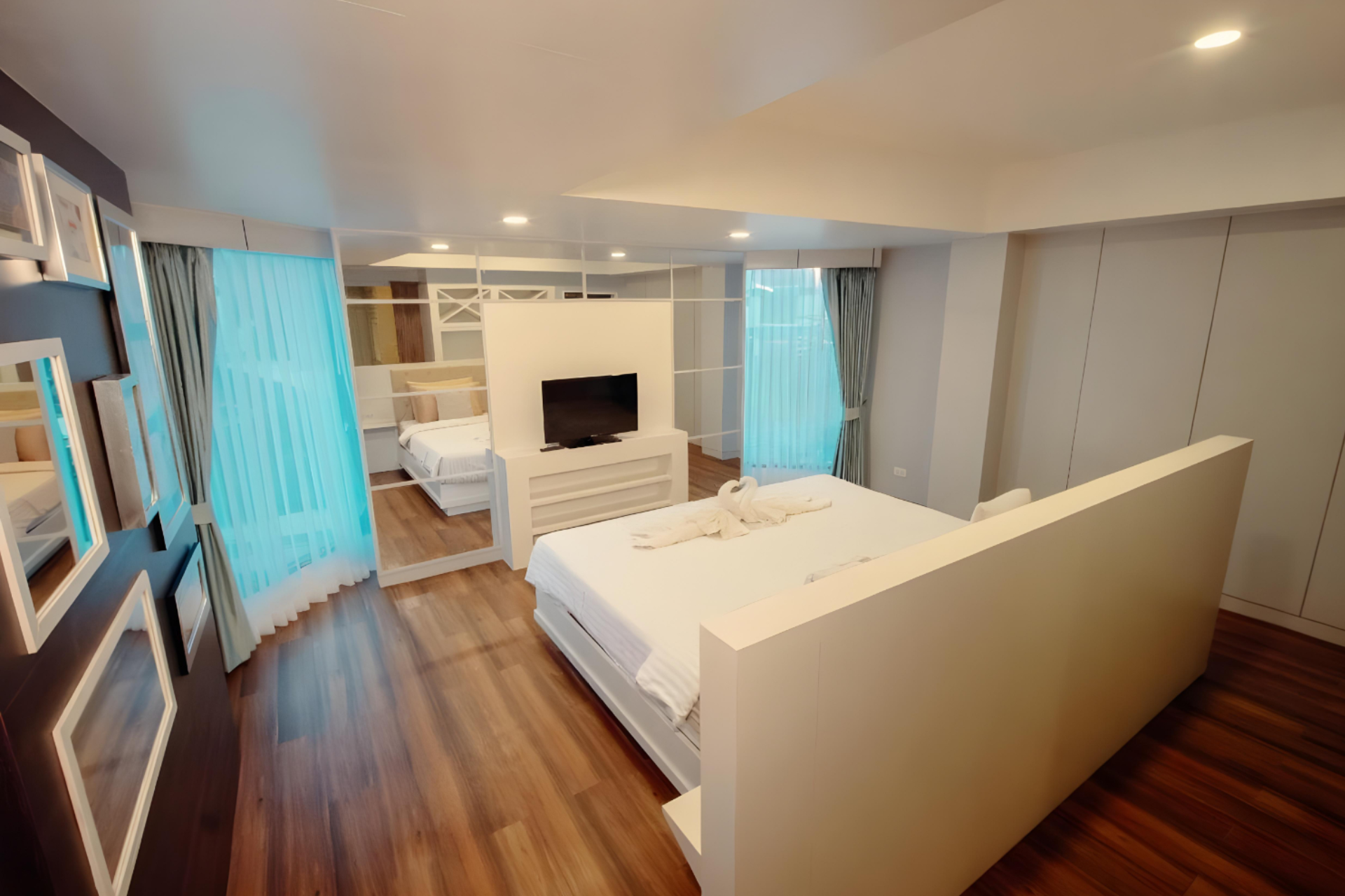 Spacious hotel room with a large white bed, towel swans, wood floors, floor-to-ceiling windows with sheer curtains, a wall-mounted TV, and mirrored walls reflecting minimalist decor.