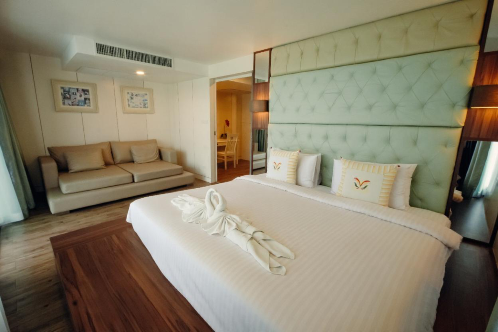 Modern bedroom with a large bed, light green tufted headboard, beige sofa, wooden floor, and towel art shaped like swans on the bed.